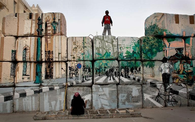 In this Tuesday, March 13, 2012 photo, a boy watches an Egyptian female artist and activist at work on the "No Walls Street" during the graffiti campaign to paint a reproduction of the streets behind them and targeted the concrete blocks walls in downtown Cairo, Egypt. After Egypt's ruling military sealed off streets around Cairo's Tahrir Square with walls of imposing concrete blocks, a group of artists decided to reopen the avenues on their own, in the public imagination, at least. (AP Photo/Nasser Nasser)