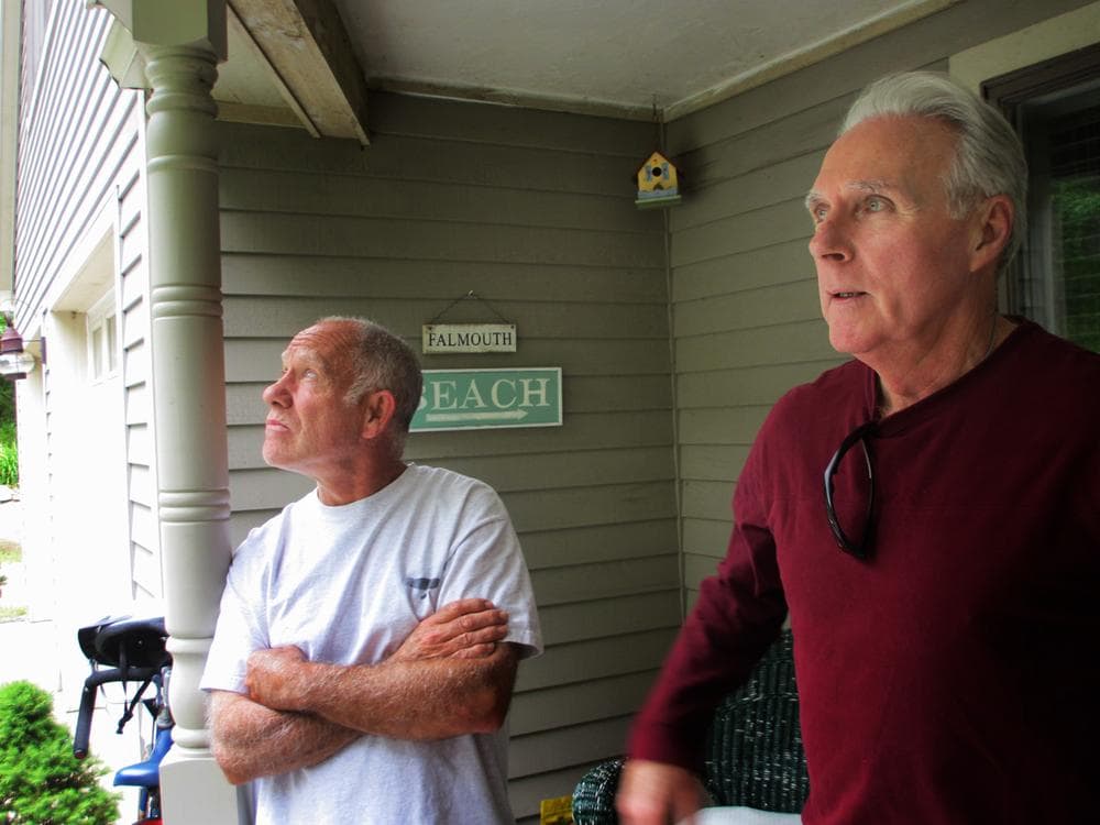Neil Andersen and John Ford stand on the back porch of Andersen’s house on Blacksmith Shop Road in Falmouth. Both say the town's wind turbines have caused them health issues. (Kathleen McNerney/WBUR)