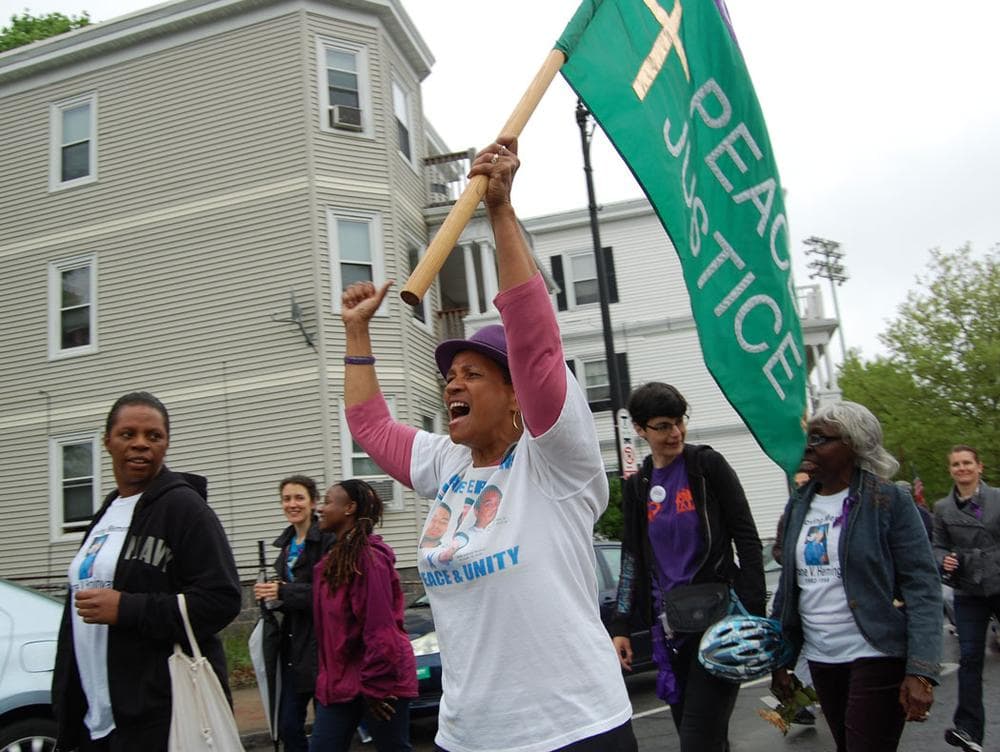 A woman wearing a shirt remembering Cape Verdean victims, including 3-year-old Malik Andrade Percival, who was fatally shot in Dorchester in 2002, waves a flag calling for “Forgiveness / Peace / Justice.” (Greg Cook)