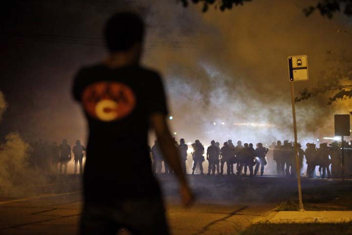 A man watches as police walk through a cloud of smoke during a clash with protesters Wednesday, Aug. 13, 2014, in Ferguson, Mo. Protests in the St. Louis suburb rocked by racial unrest since a white police officer shot an unarmed black teenager to death turned violent Wednesday night, with people lobbing Molotov cocktails at police who responded with smoke bombs and tear gas to disperse the crowd. (AP)