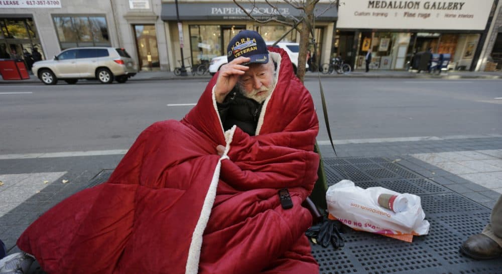 A homeless man adjusts his hat while wrapped in a blanket on a sidewalk on Boylston Street in Boston. In the year since the Long Island Bridge closure cut off access to a homeless shelter on the island, homelessness has risen 5.6 percent in Boston. (Steven Senne/AP)