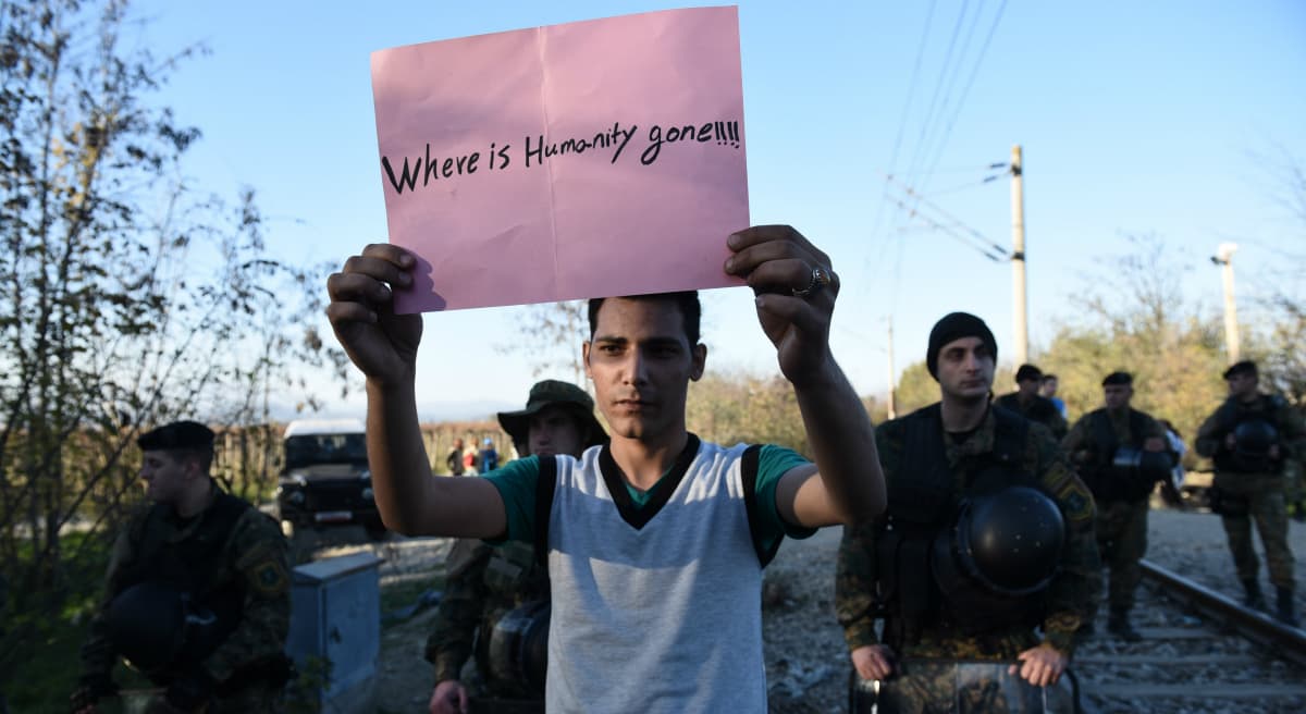 A migrant holds a sign reading ''Where is humanity gone'' at no man's land, as Macedonian policemen guard the borders of their country near Idomeni village, northern Greece on Thursday, Nov. 19, 2015. (Giannis Papanikos/ AP) 