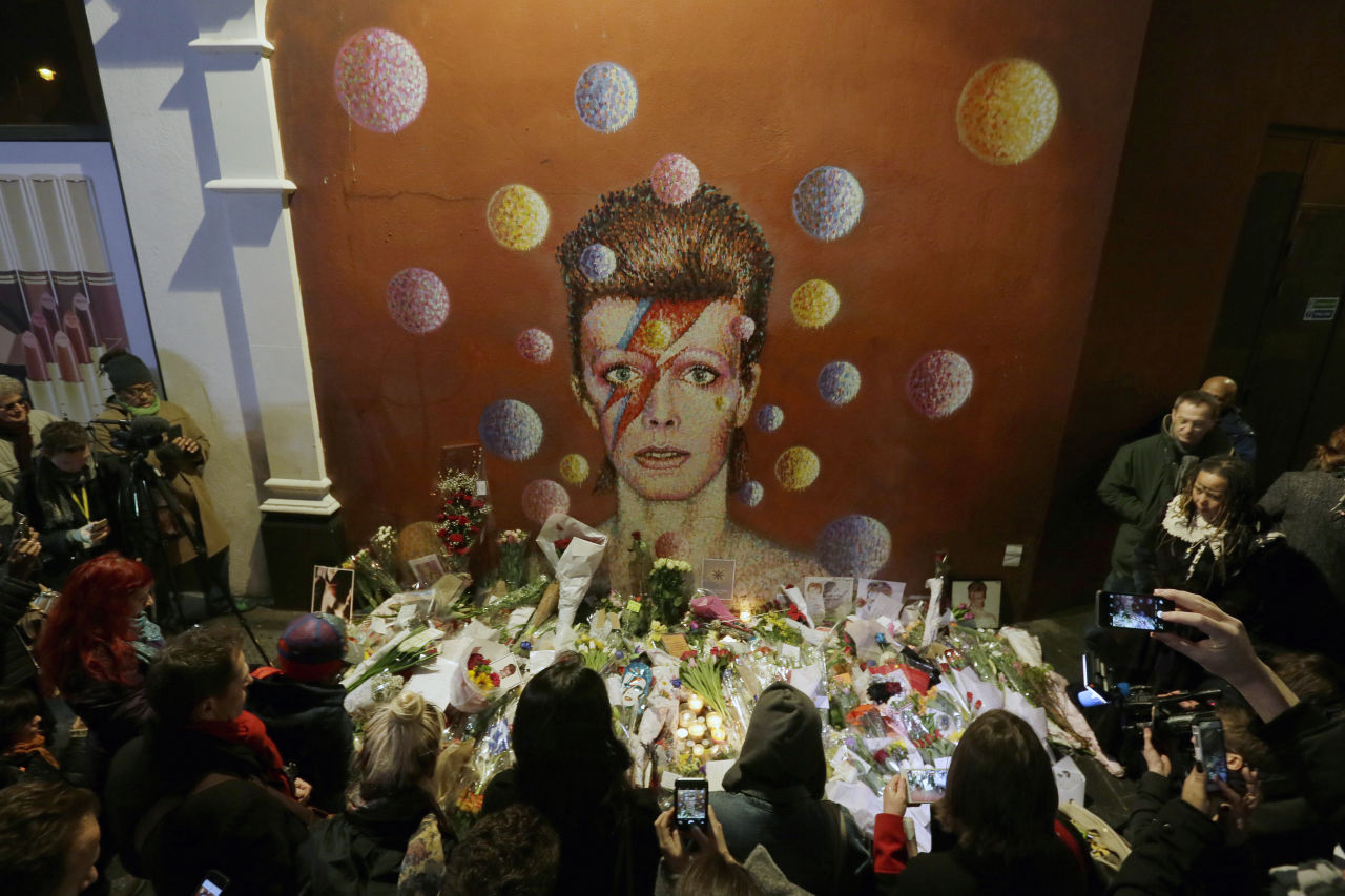 People gather next to tributes placed near a mural of British singer David Bowie by artist Jimmy C, in Brixton, south London on Monday, Jan. 11, 2016. Bowie, the other-worldly musician who broke pop and rock boundaries with his creative musicianship, nonconformity, striking visuals and a genre-spanning persona he christened Ziggy Stardust, died of cancer Sunday aged 69. He was born in Brixton. (Tim Ireland/AP)