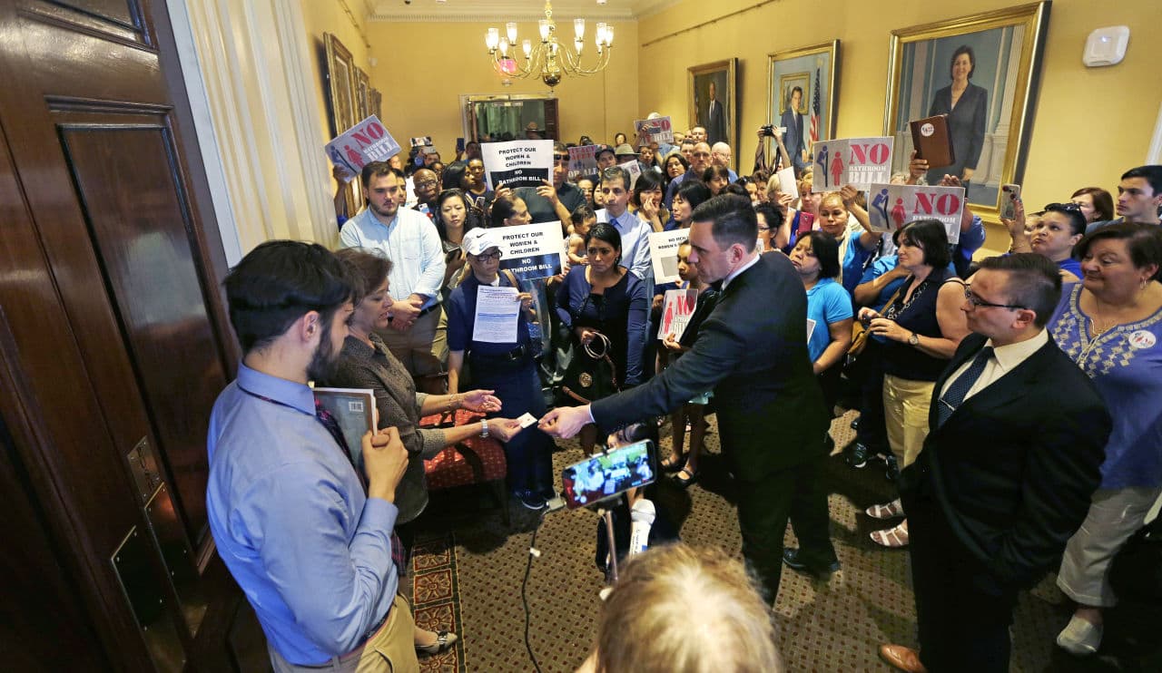 Andrew Beckwith, president of Massachusetts Family Institute, hands his business card to members of Governor Baker&#039;s staff, who block the door to his office, as protesters gather at the State House. (Charles Krupa/AP)