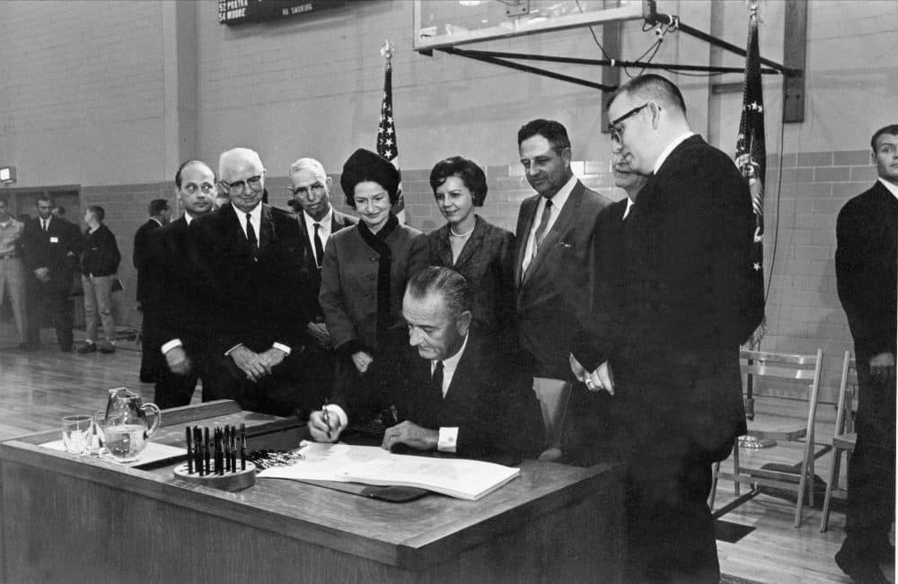 U.S. President Lyndon B. Johnson, seated at the desk he used while a student secretary at Southwest Texas State College 35 years ago, signs the Higher Education Act at San Marcos, Texas, Nov. 8, 1965.  Looking over the president's shoulder is Lady Bird Johnson and to her right are Mrs. Jake Pickle; Rep. James Jarrell &quot;Jake&quot; Pickle, D-Texas; J.C. Kellam, of Austin, partially hidden; and James H. McCrocklin, president of the college.  The men at extreme left are not identified.  (AP Photo)