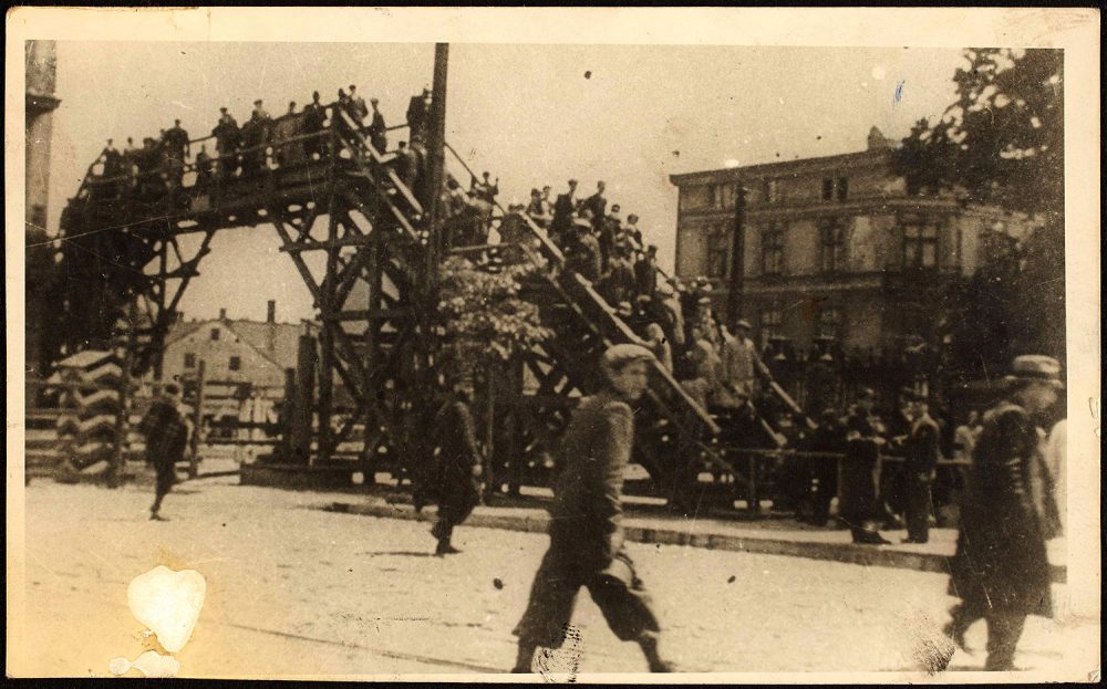 Henryk Ross photo of a boy walking in front of a Lodz Ghetto bridge at Zigerska Street, with residents crossing, c. 1940-1944. (Courtesy, Museum of Fine Arts, Boston)