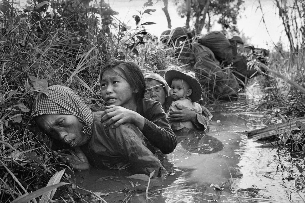 Women and children crouch in a muddy canal as they take cover from intense Viet Cong fire, January 1, 1966. Paratroopers of the 173rd Airborne Brigade (background) escorted the civilians through a series of firefights during the U.S. assault on a Viet Cong stronghold at Bao Trai, about twenty miles west of Saigon. (Horst Faas/AP)