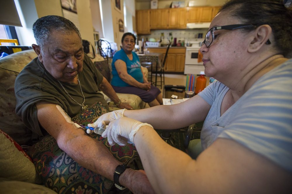 Fausto Alvarado looks down while his daughter, Cruz Romero, administers an intravenous injection of an antibiotic to treat his lung infection. Alvarado's medical condition does not permit him to have an air-conditioner, which during the summer months makes breathing difficult. (Jesse Costa/WBUR)