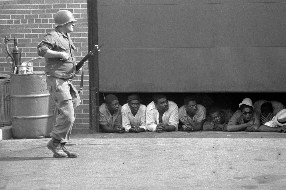 In this July 26, 1967 file photo, an Army soldier stands guard as men captured in the vicinity of the 10th Police Precinct in Detroit peer from under a garage door awaiting transfer. Five days of violence would leave 33 blacks and 10 whites dead, and more than 1,400 buildings burned. More than 7,000 people were arrested. (AP)