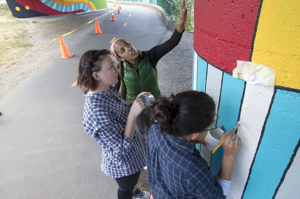Artist Silvia López Chavez discusses the colors she wants for the pillar with volunteer artist Katie Miller as artist Sara Barrientos continues her work on the wall. (Jesse Costa/WBUR)