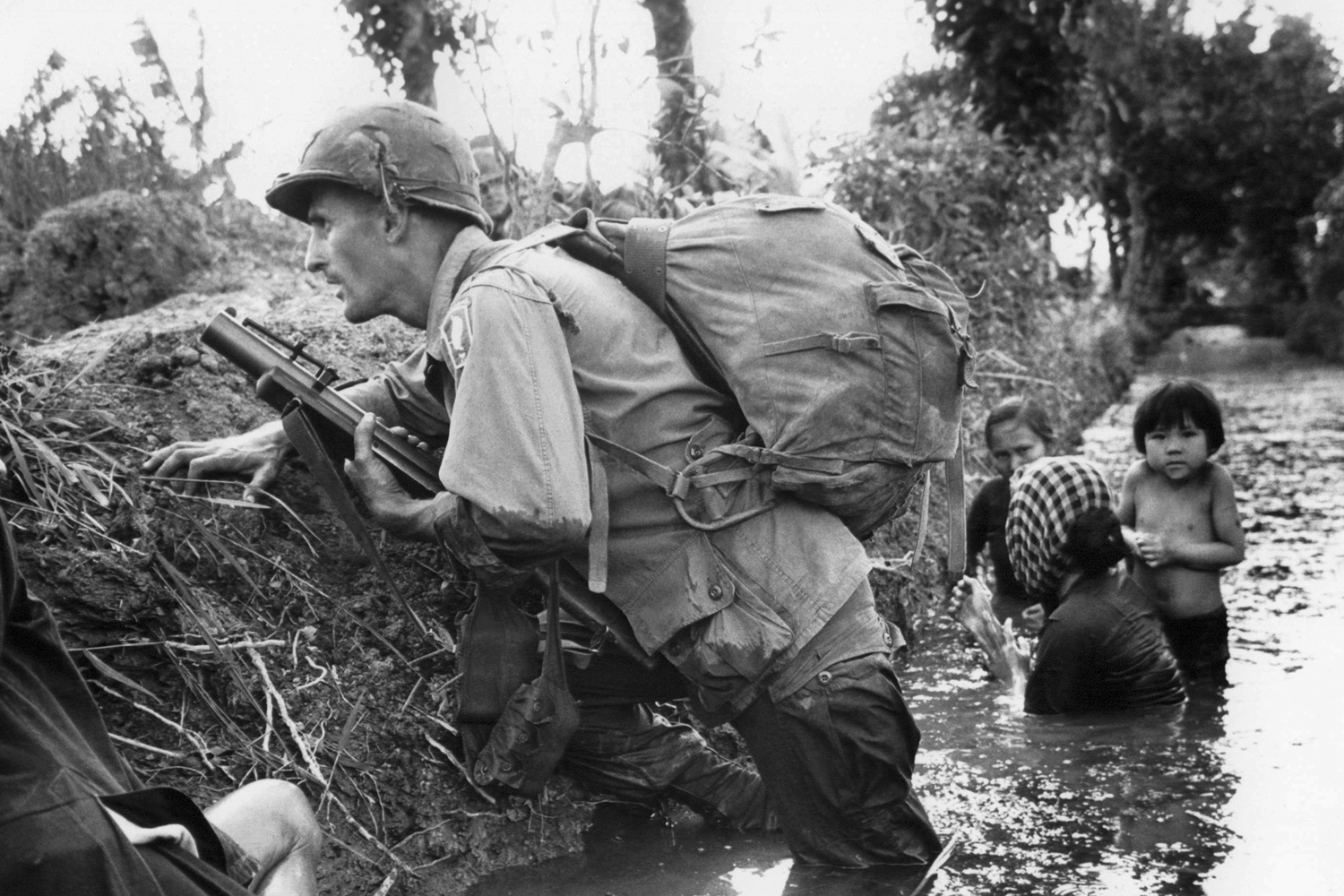 In this Jan. 1, 1966 file photo, a Paratrooper of the 173rd U.S. Airborne brigade crouches with women and children in a muddy canal as intense Viet Cong sniper fire temporarily pins down his unit during the Vietnamese War near Bao trai in Vietnam. (Horst Faas/AP)