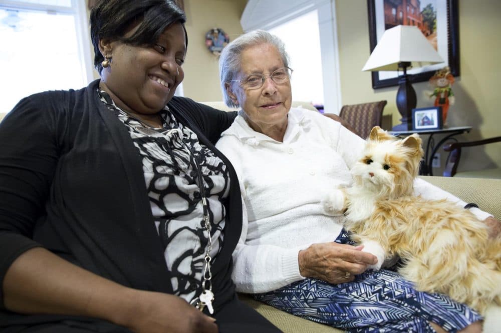 Caretaker Mirlene Desrosiers sits with Lillian Hall and Milo the robotic cat at the Brookdale Senior Living Community in Danvers. (Robin Lubbock/WBUR)