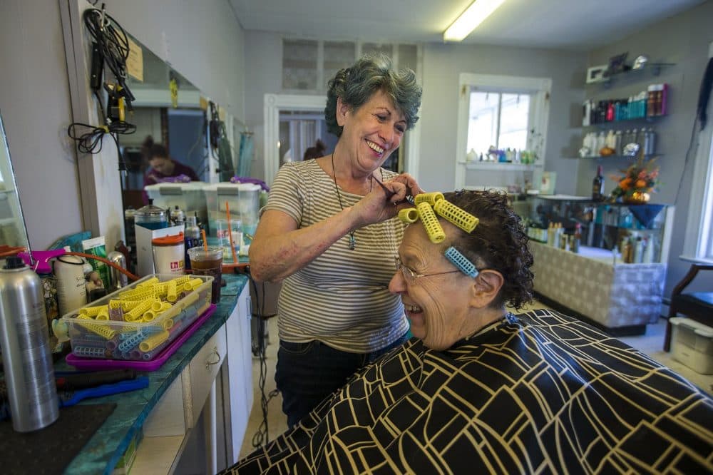 Judy Wilkins calls herself the poster child for a new type of cancertherapy called CAR-T. As she is having a laugh, Wilkins is putting hair rollers into Annina McCully's hair at her salon in Woburn. (Jesse Costa/WBUR)