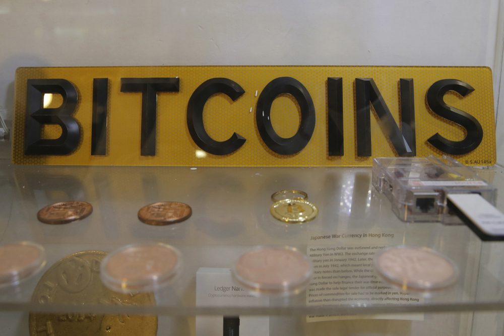 In this Friday, Dec. 8, 2017, photo, coins and sign are displayed next to a Bitcoin ATM in Hong Kong. The launch of a U.S. futures contract for bitcoin on Sunday, Dec. 10, 2017, underscores the virtual currency's increasing mainstream acceptance, including in many parts of Asia, where it already has a wide following among speculators and investors. (AP Photo/Kin Cheung)