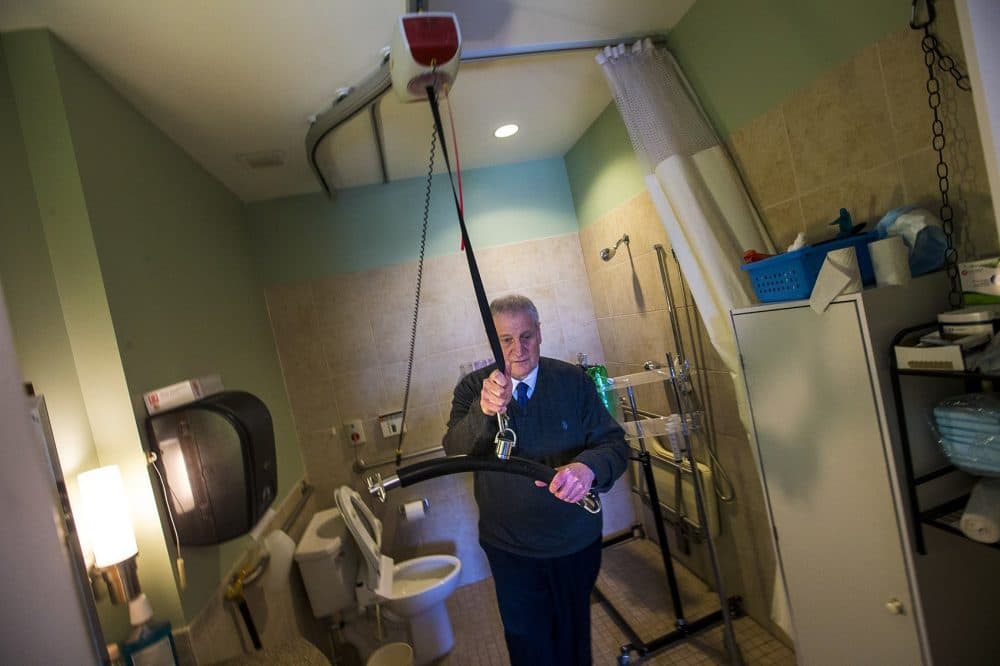 Barry Berman, CEO of Chelsea Jewish Lifecare, which operates the Leonard Florence Center For Living, demonstrates the lift that aides use to get Steve from his bed into the bathroom. (Jesse Costa/WBUR)