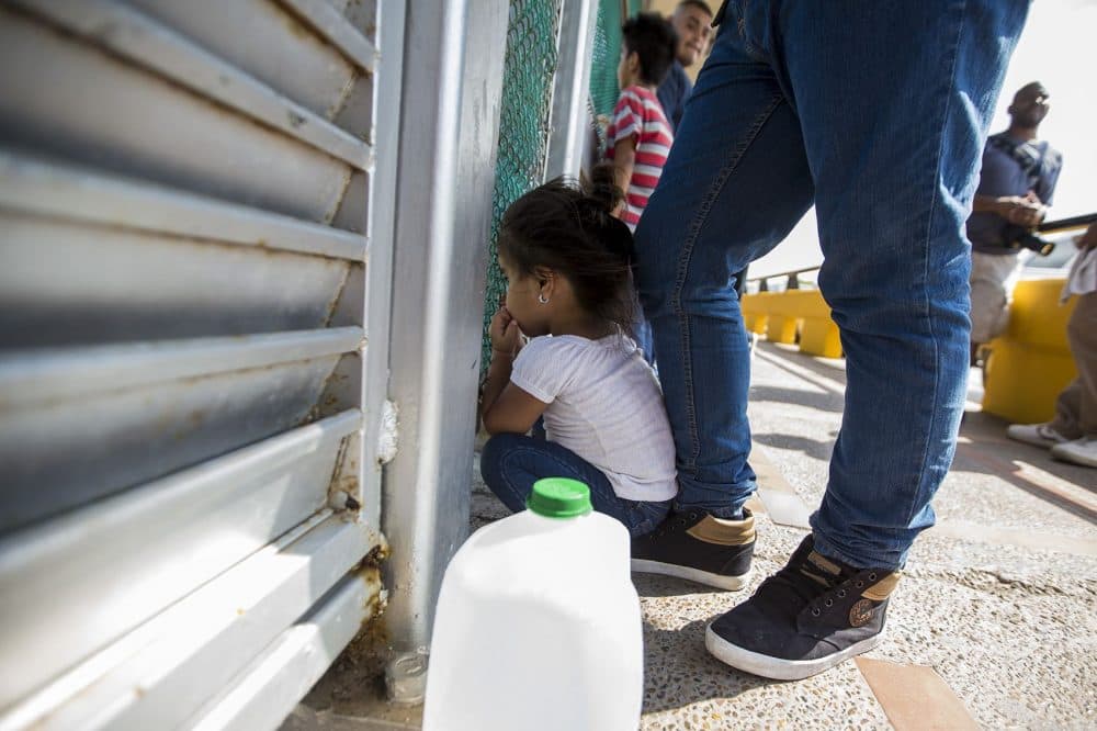 A 3-year-old from Honduras peers through a fence at the U.S.-Mexico border while her family waits to apply for asylum. (Jesse Costa/WBUR)