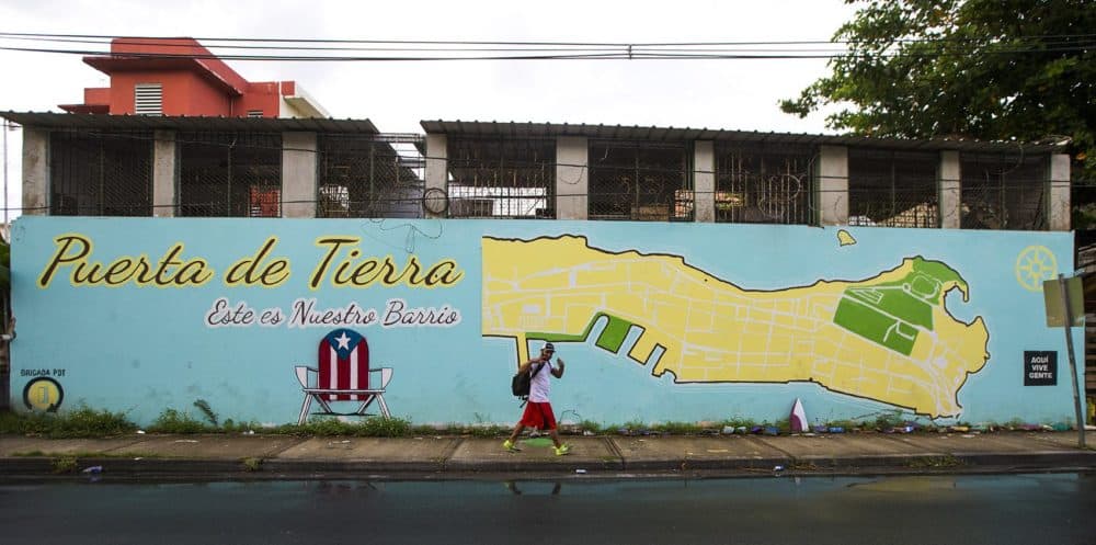 A man flashes peace signs as he walks past a mural in Puetra de Tierra, one of the poorest areas in San Juan. Community organizers are working to revitalize the neighborhood with an outreach campaign that includes the slogan ‘AQUÍ VIVE GENTE’ - 'WE LIVE HERE.' (Jesse Costa/WBUR)