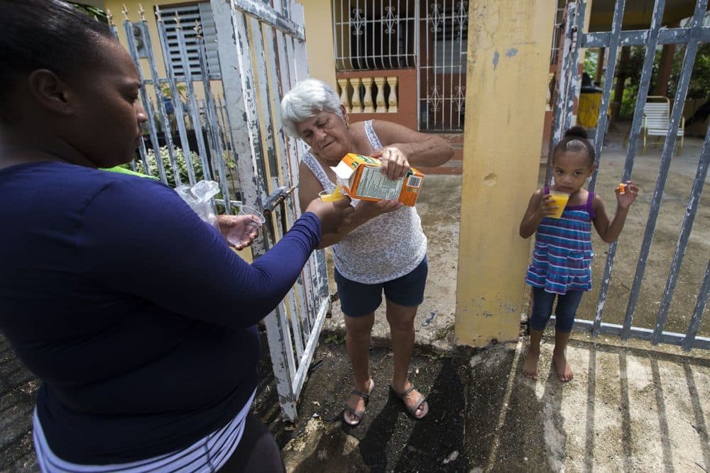 Juana Alicea sirve jugo de naranja en tazas para Indiana Medina y para Kendra, de 3 años de edad, cuando pasan frente a su casa en un día cálido de Toa Baja. (Jesse Costa/WBUR)