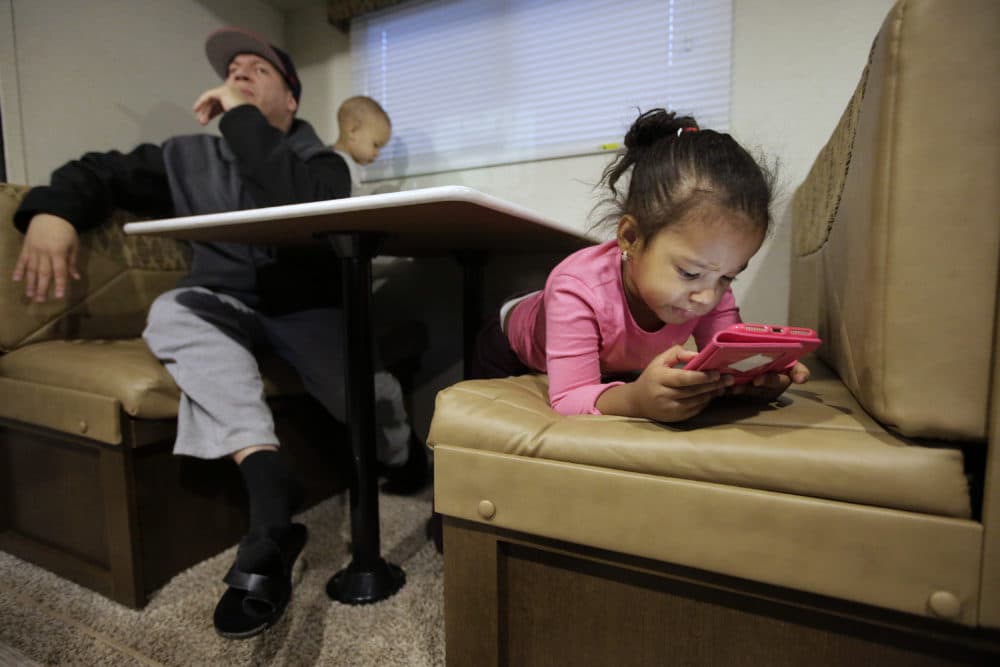 Four-year-old Isairis Grullon, right, looks at a mobile device as her father Jose Grullon, 45, and her 1-year-old brother Jadiel Grullon are seated at a table in a trailer home at a trailer camp in Lawrence for people displaced by gas line failures and explosions. (Steven Senne/AP)
