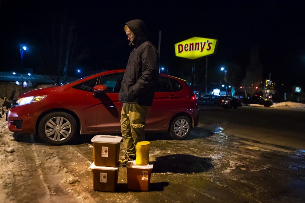 In a Denny's parking lot in Auburn, Maine, Church of Safe Injection founder Jesse Harvey stands behind four containers filled with used needles collected from drug users around Lewiston throughout the evening. (Jesse Costa/WBUR)