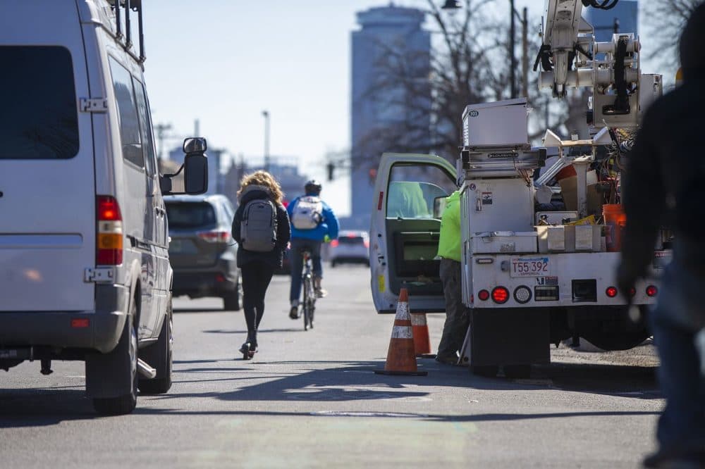 A cycylist and a young woman on a scooter manuver between open doors of double-parked vehicles and the flow of traffic down Commonwealth Ave. (Jesse Costa/WBUR)