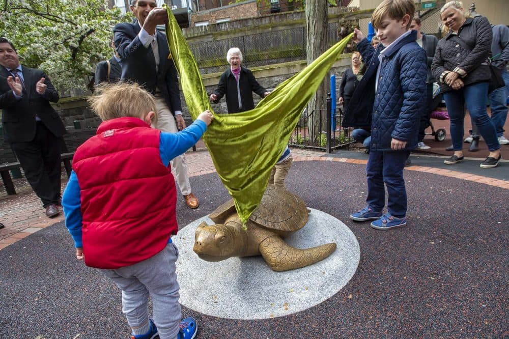 Sculptor Nancy Schön cheers from behind as Miguel Rosales, left, and three young helpers unveil the bronze recreation of “Myrtle The Turtle,” the famed green sea turtle of the New England Aquarium, at the Myrtle Street Playground on Beacon Hill. (Jesse Costa/WBUR)