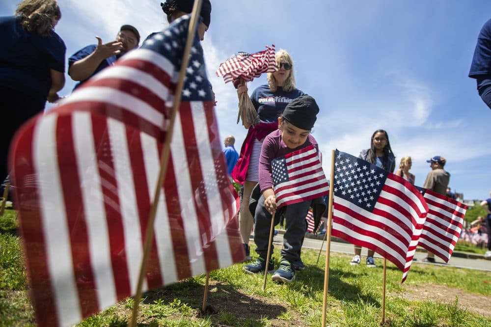 With the help of Kathy Sherman, holding the flags in the background, Paramvir Saini sticks an American flag into the ground. (Jesse Costa/WBUR)