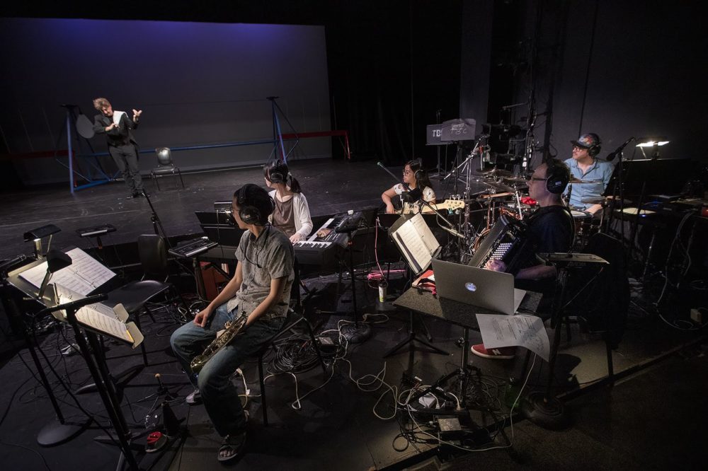 The ensemble awaits to play along as comedian Joel Jeske begins to motion to play a violin. (Jesse Costa/WBUR)