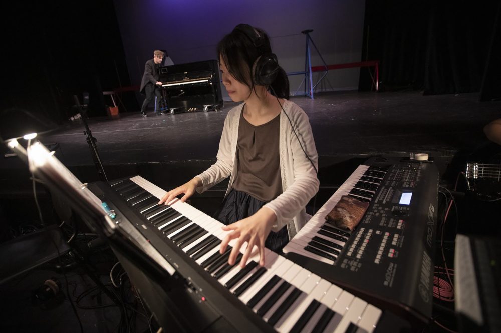 Student composer/pianist Yoko Suzuki playing as Joel Jeske emerges onto the stage pushing his piano. (Jesse Costa/WBUR)