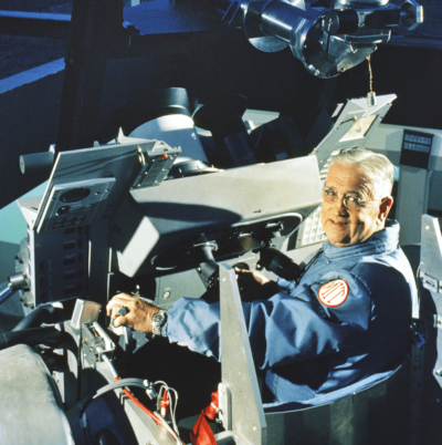 Dr. Charles “Doc” Stark Draper sits in a blue jumpsuit inside the simulator. On his should is an &quot;MIT&quot; patch.
