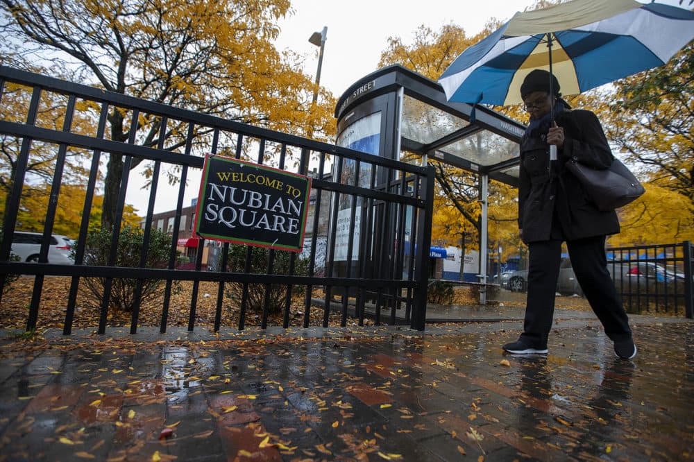 A woman walks past a “Welcome to Nubian Square” sign at the gate of the Dudley Square Plaza on Washington St. (Jesse Costa/WBUR)