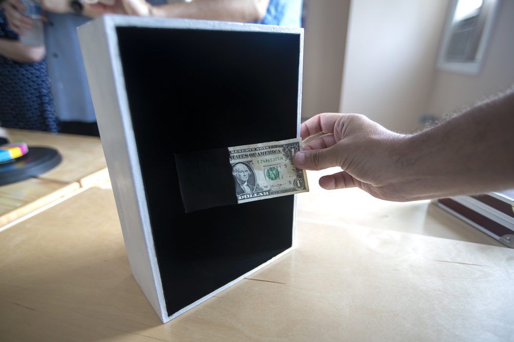 A $1 bill, partially painted with the Singularity Black coating, demonstrates it’s ability to absorb 99% of the light when placed into a box painted with the same coating. (Jesse Costa/WBUR)
