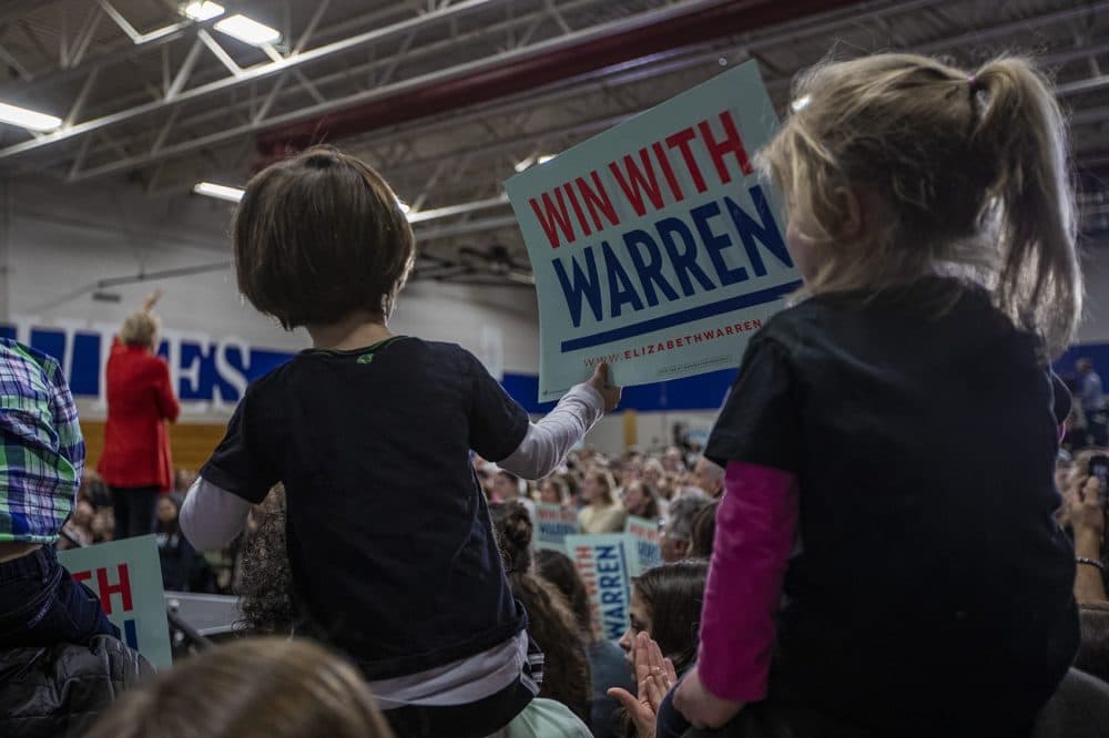 Two young children sitting on the shoulders of their parents wave a Warren campaign sign during the candidate's speech at Rundlett Middle School in Concord, NH. (Jesse Costa/WBUR)