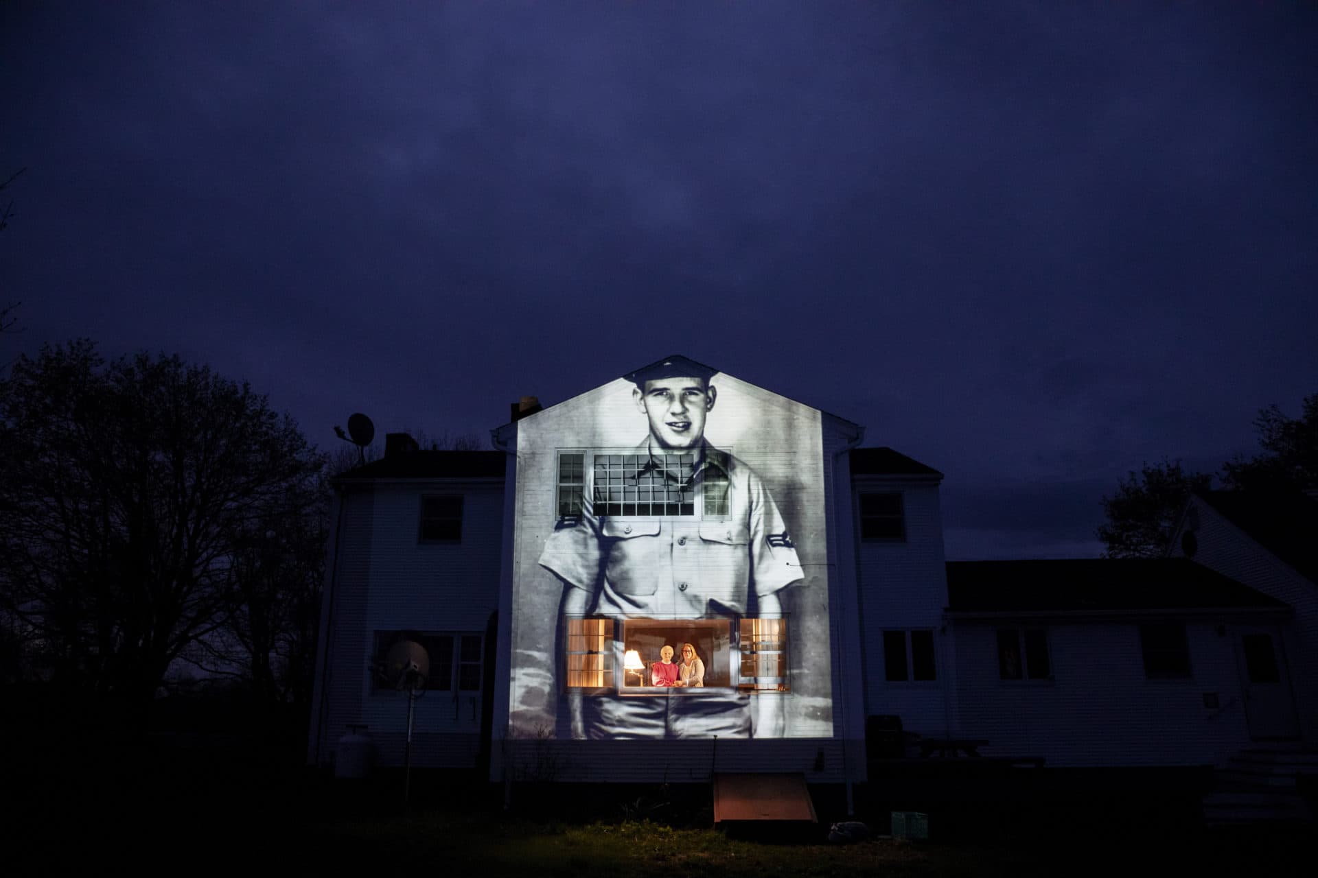 An image of veteran Charles Lowell is projected onto the home he shared with his wife, Alice, for 30 years as she stands at left with her daughter, Susan Kenney, in Hardwick, Mass. (David Goldman/AP)