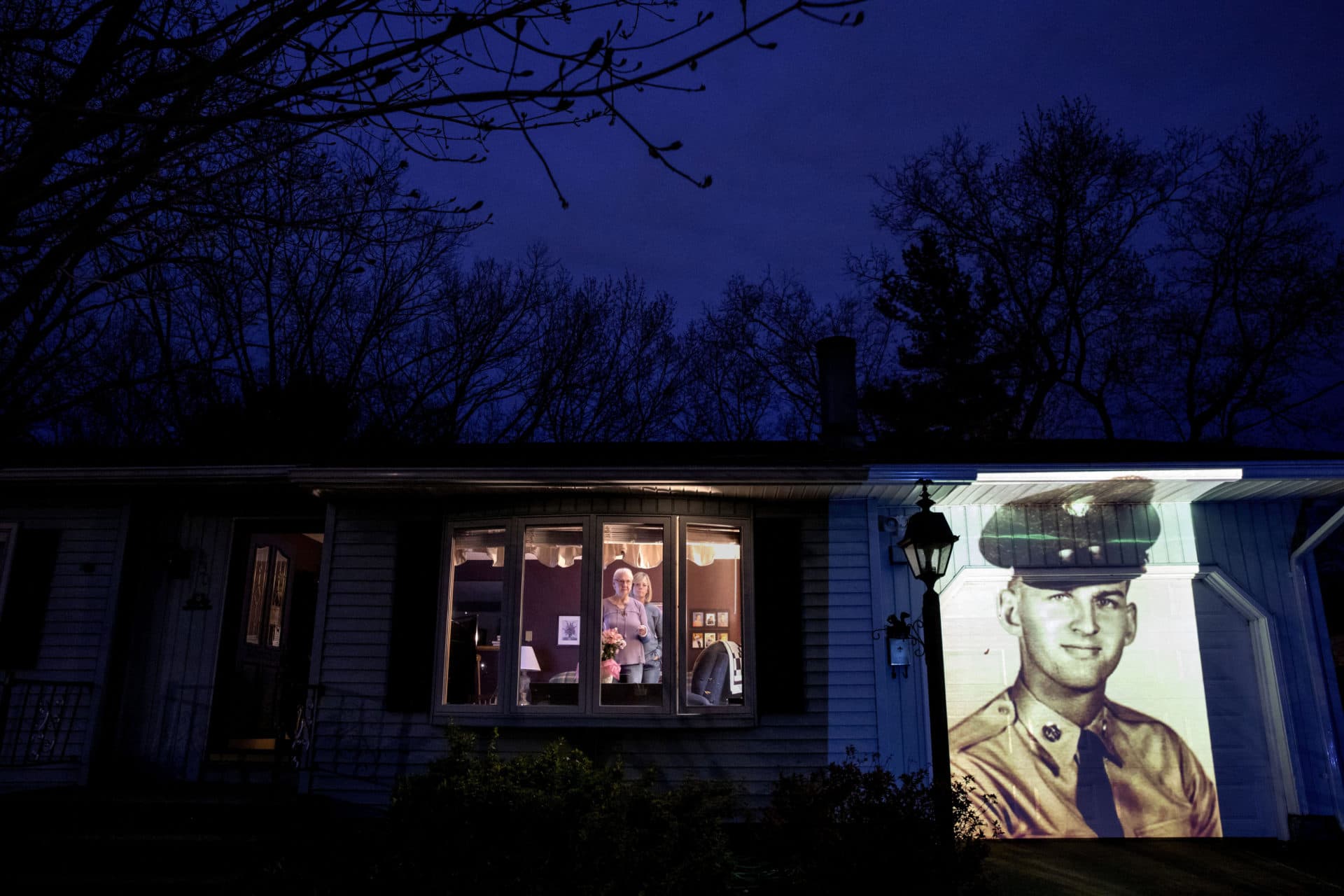An image of veteran Francis Foley is projected onto the home of his wife, Dale Foley, left, as she looks out a window with their daughter, Keri Rutherford, in Chicopee, Mass. (David Goldman/AP)