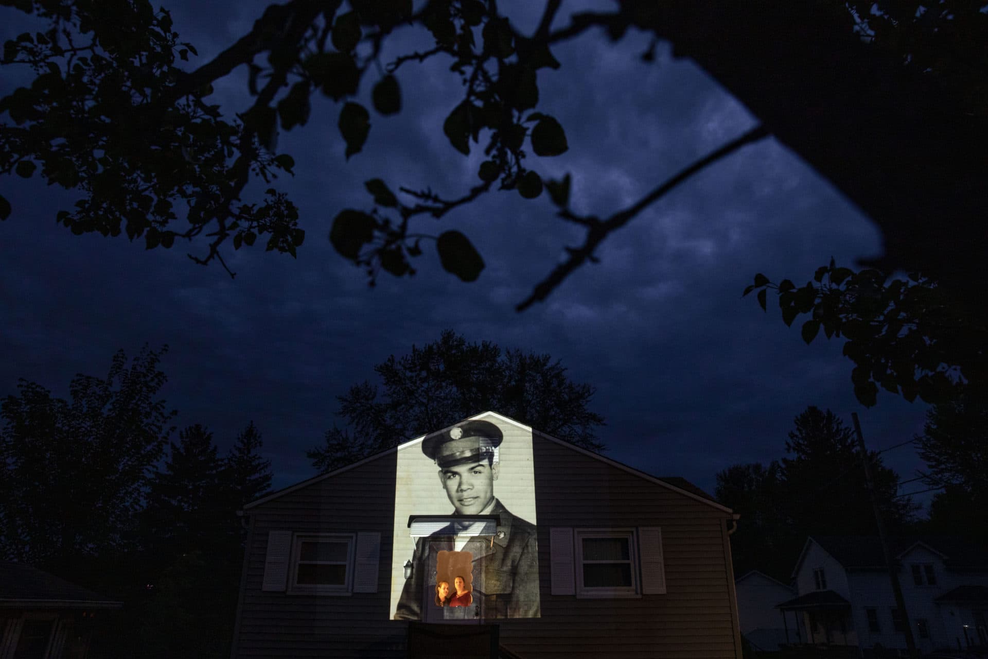An image of veteran Samuel Melendez is projected onto the home of his nieces, Janet Ramirez, right, and Mary Perez, as they look out a doorway in Chicopee, Mass. (David Goldman/AP)
