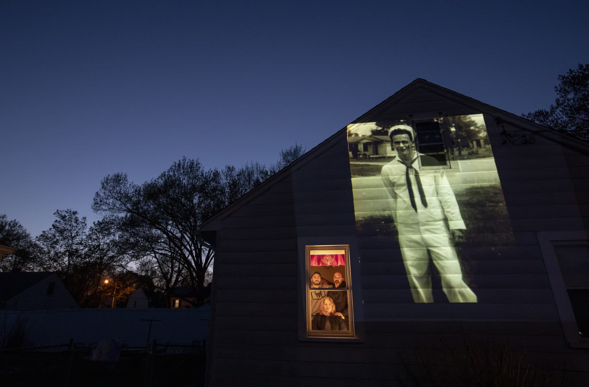 An image of veteran James Mandeville is projected onto the home of his daughter, Laurie Mandeville Beaudette, as she looks out a window with her son, Kyle, left, and husband, Mike, in Springfield, Mass. (David Goldman/AP)