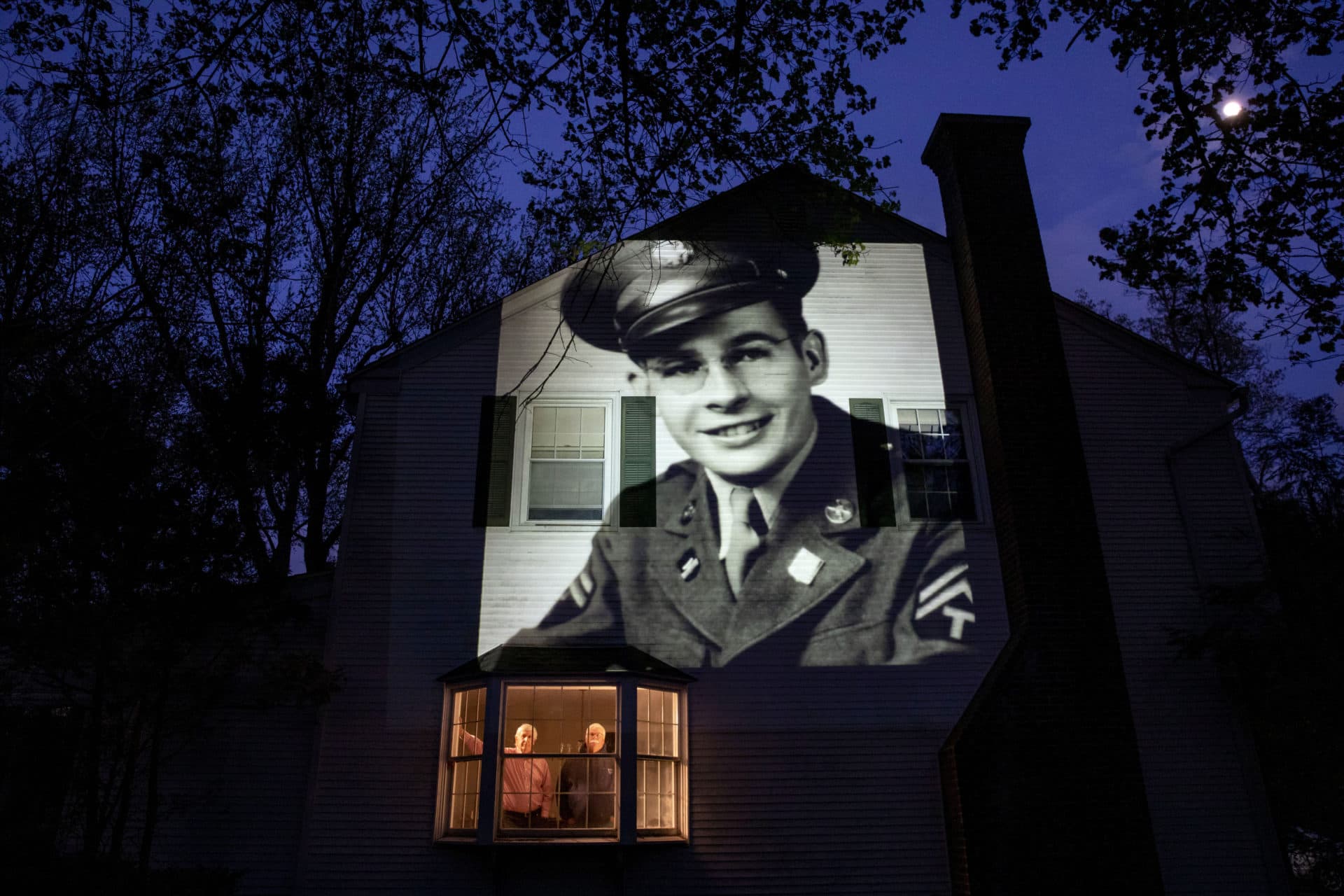 An image of veteran James Sullivan is projected onto the home of his son, Tom Sullivan, left, as he looks out a window with his brother, Joseph Sullivan, in South Hadley, Mass. (David Goldman/AP)