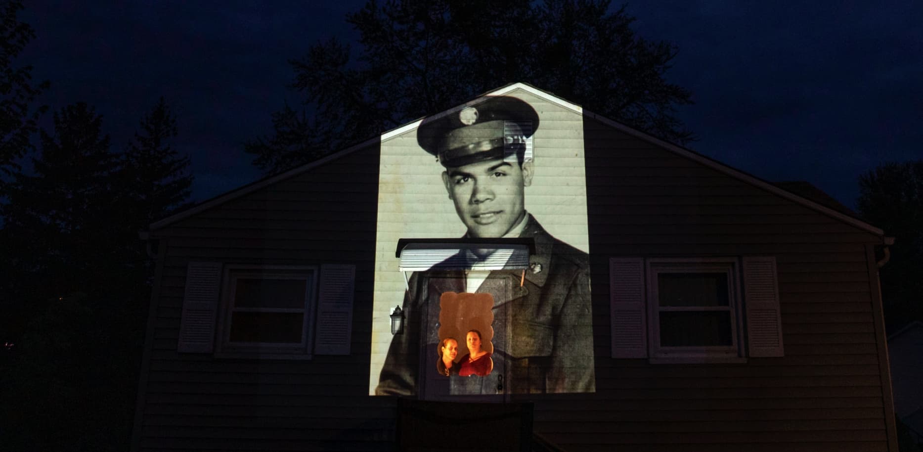 An image of veteran Samuel Melendez is projected onto the home of his nieces, Janet Ramirez, right, and Mary Perez, as they look out a doorway in Chicopee, Mass. (David Goldman/AP)