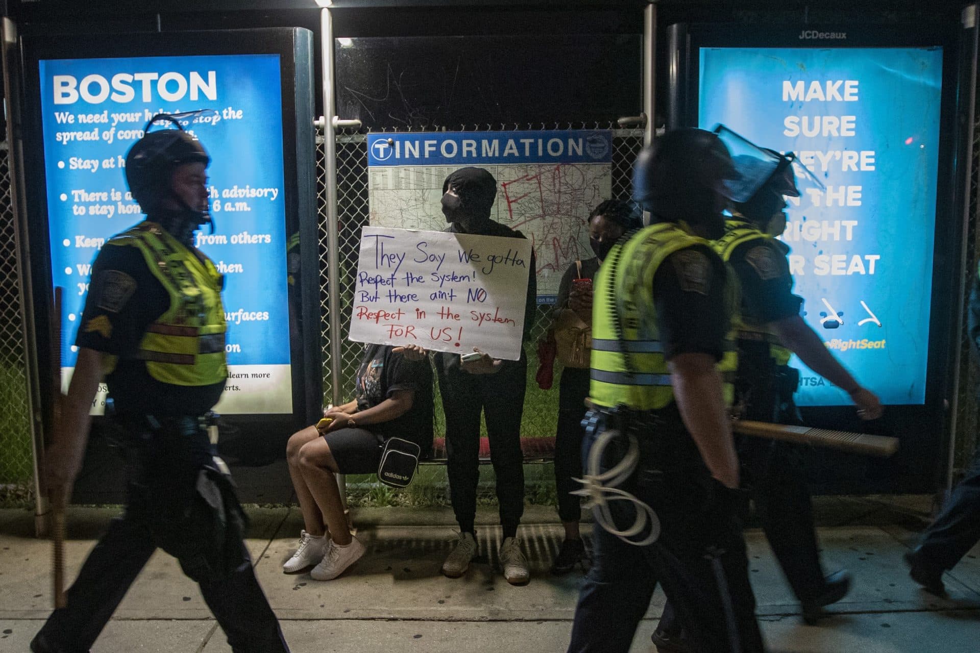 Boston Police walk past protesters waiting in a bus stop on Washington Street as they walk to the District E-13 station in Jamaica Plain. (Jesse Costa/WBUR)