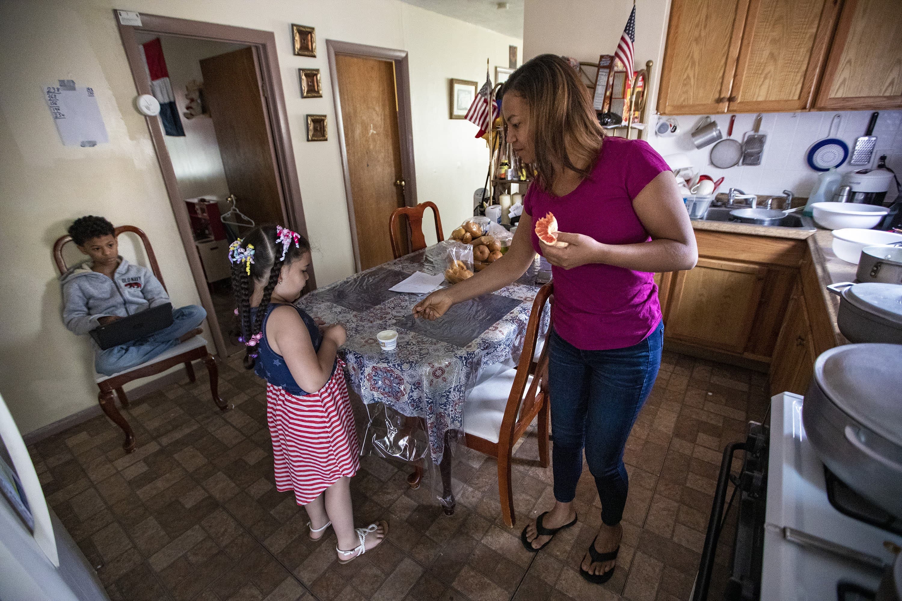 Margareth le da una cuchara a Lucero para que coma su bocadillo de yogur a última hora de la mañana, mientras Jesús se sienta en una silla usando una computadora portátil. (Jesse Costa / WBUR)