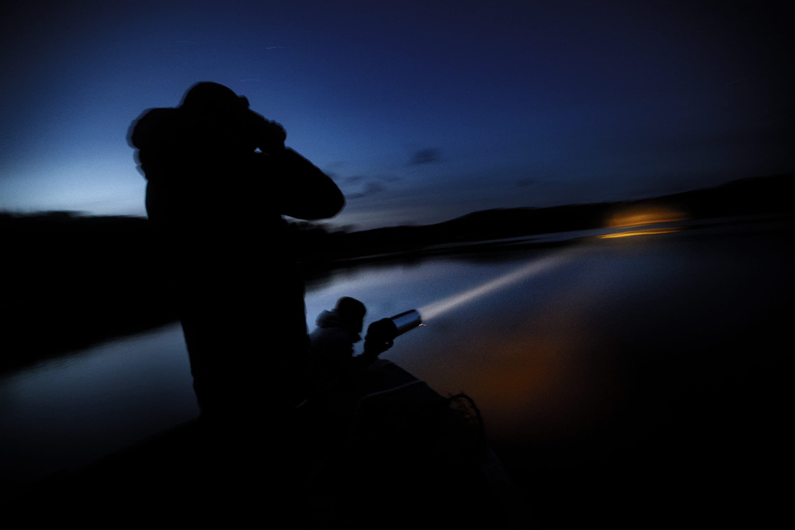 Mark Burton shines a spotlight across the water as Chris Persico uses a pair of binoculars to find loon chicks on Rangeley Lake in Rangeley, Maine. (Jesse Costa/WBUR)