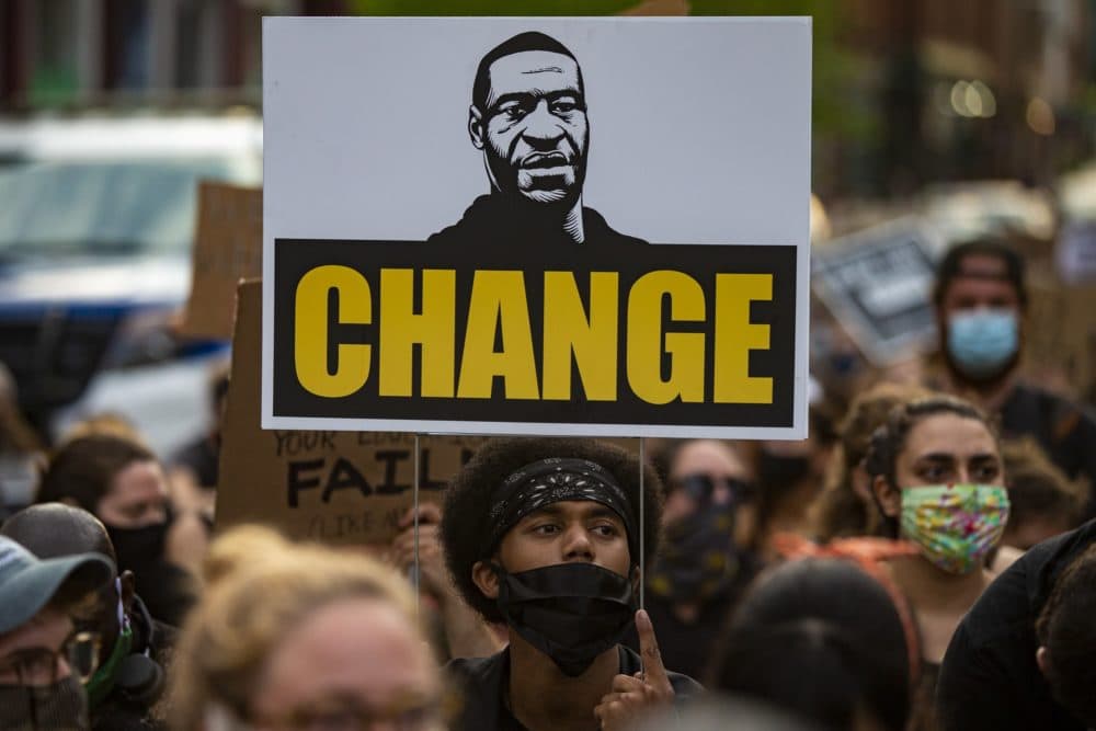 A protester sits and holds a sign with George Floyd’s image during an 8 minute and 26 second moment of silence in front of the Boston police substation in Jamaica Plain in 2020. (Jesse Costa/WBUR)