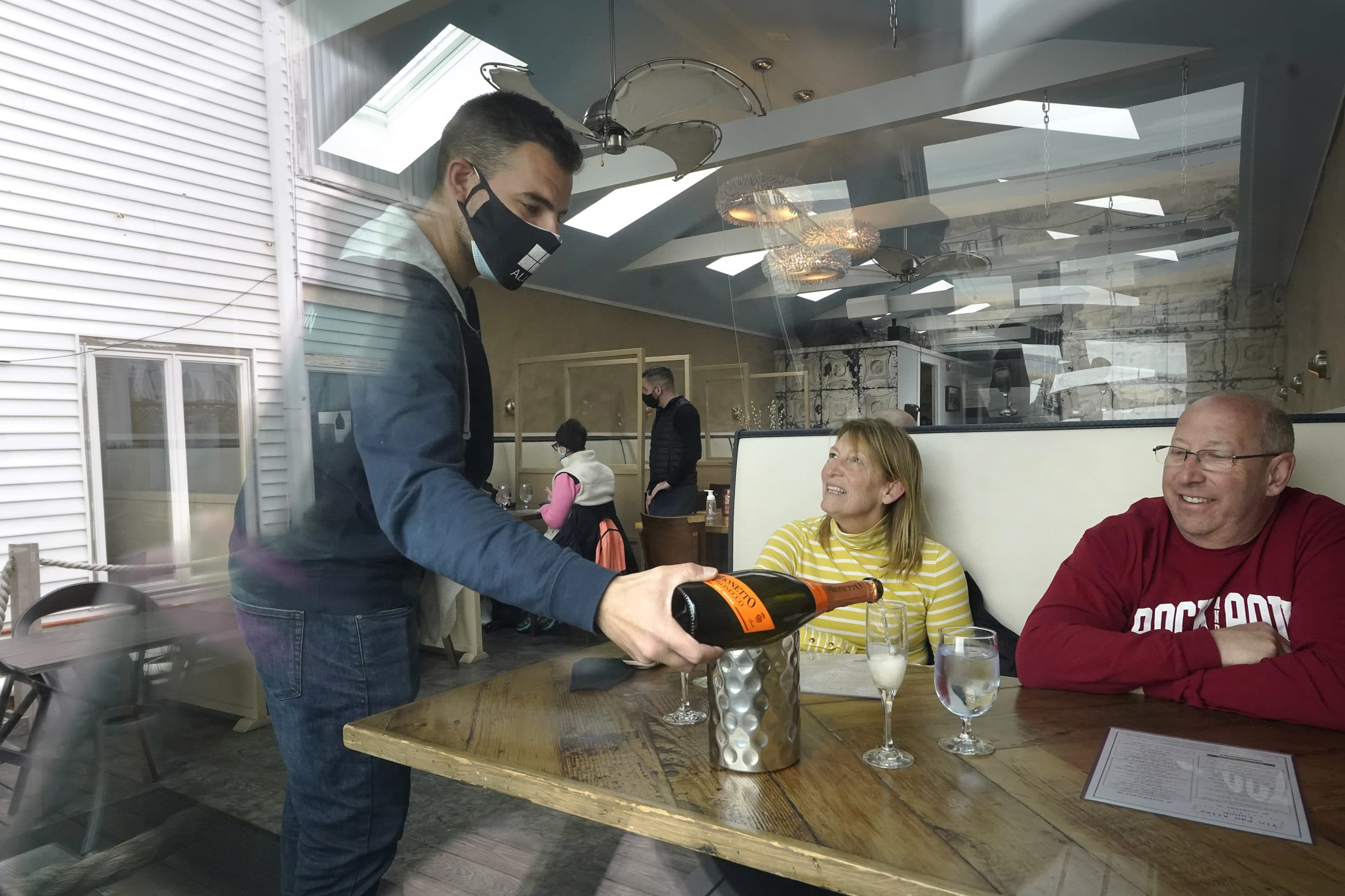 Bartender Denis Angelov, of Provincetown, Mass., left, serves sparking wine to Julie Skaller, left, and her husband David Skaller, right, both of Brewster, N.Y., at Tin Pan Alley restaurant, April 6, 2021, in Provincetown. Hotels, restaurants and other businesses in tourist destinations are warning that hiring challenges during the coronavirus pandemic could force them to pare back operating hours or curtail services just as they’re eyeing a bounce-back summer. (Steven Senne/AP)