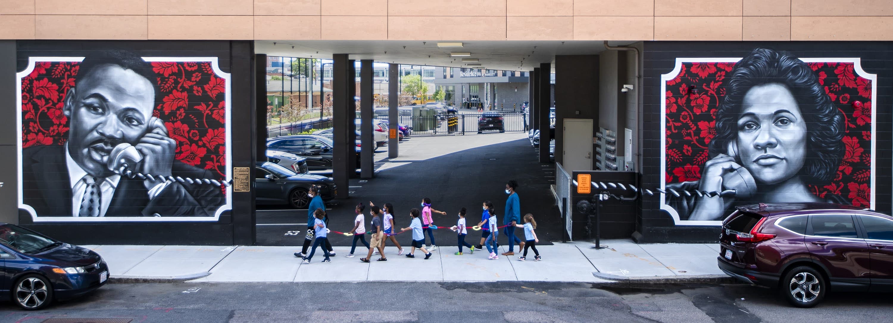 Young school children and their teachers pass the ‘Roxbury Love Story’ mural by ‘Problak’ Gibbs, depicting Rev. Martin Luther King, Jr. and Coretta Scott King speaking to each other through corded telephones on either side of the archway. The mural is on an exterior wall of an apartment building in the former location of the Twelfth Baptist Church, where King served as assistant minister from 1951 to 1954. (Jesse Costa/WBUR)