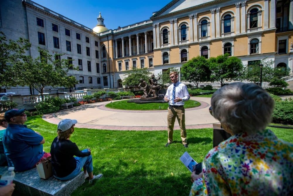 A small group of State House tour participants sit in the shade during a hot sunny day  as they listen to Suffolk University. senior Alex Svenson explain the origin of the Massachusetts Firefighters Memorial behind him. (Jesse Costa/WBUR)