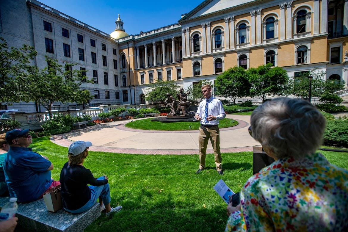 A small group of State House tour participants sit in the shade during a hot sunny day as they listen to Suffolk University. senior Alex Svenson explain the origin of the Massachusetts Firefighters Memorial behind him. (Jesse Costa/WBUR)