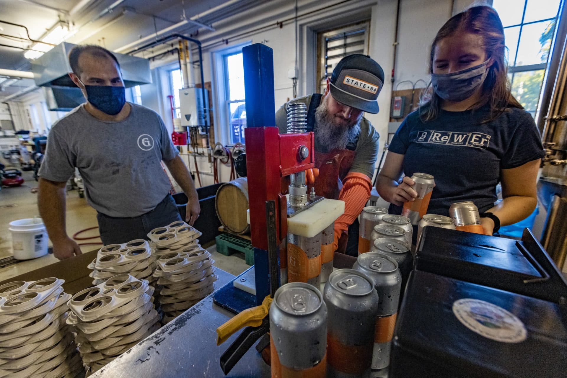 Paul Gentile and Caroline LeBlanc of Gentile Brewing in Beverly watch as State 64 owner Tim Little makes an adjustment to a beer carrier topper; using a traditional bottle capper fitted with a specially designed block to press down the the E6PR carriers on top of cans more efficiently. (Jesse Costa/WBUR)