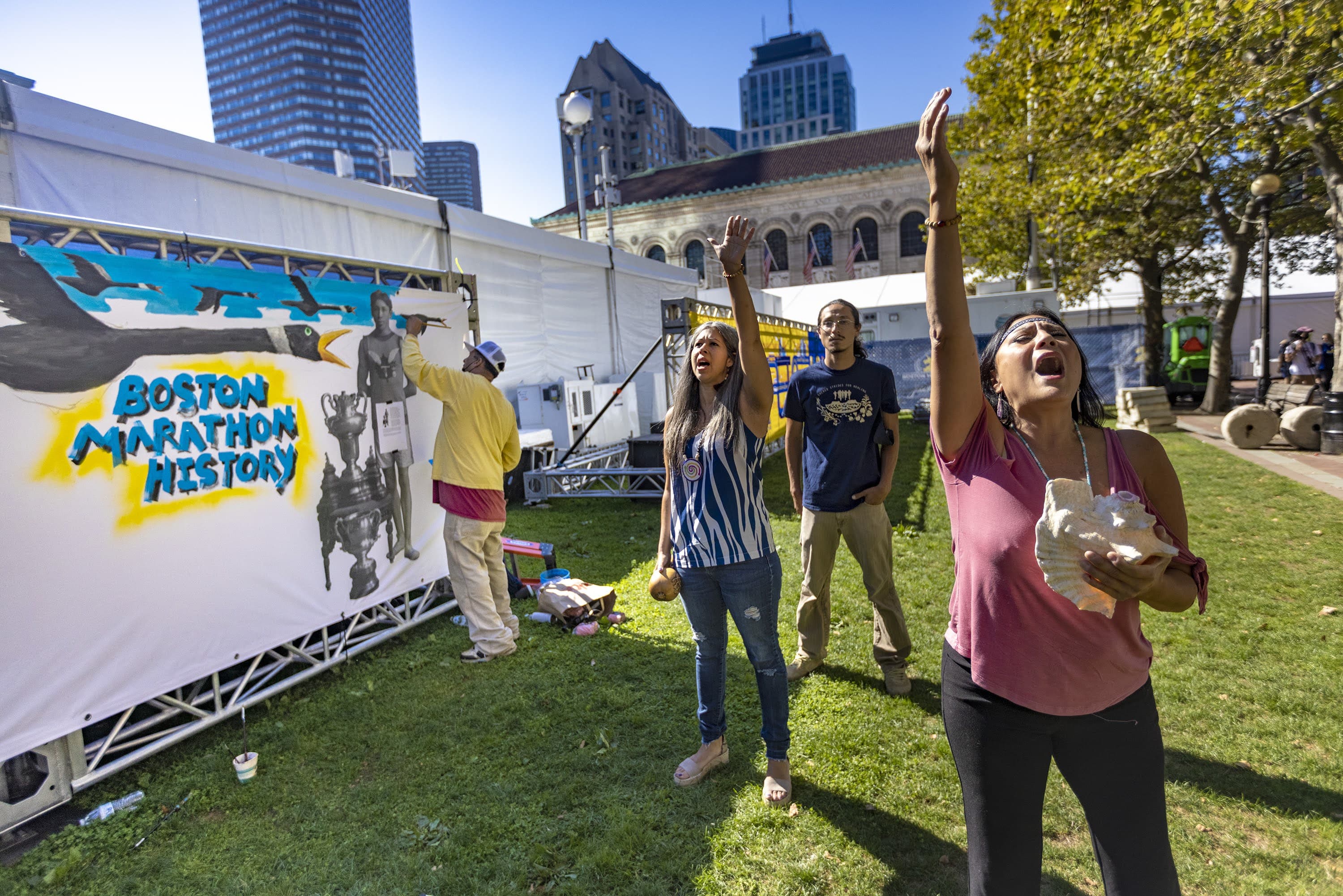 Chali’Naru Dones, right, and Darlene Flores, co-founders of the Indigenous Peoples' Day Committee in Newton, perform a healing song as artist Robert Peters paints a mural illustrating Indigenous history in the Boston Marathon near the finish line in Copley Square. (Jesse Costa/WBUR)