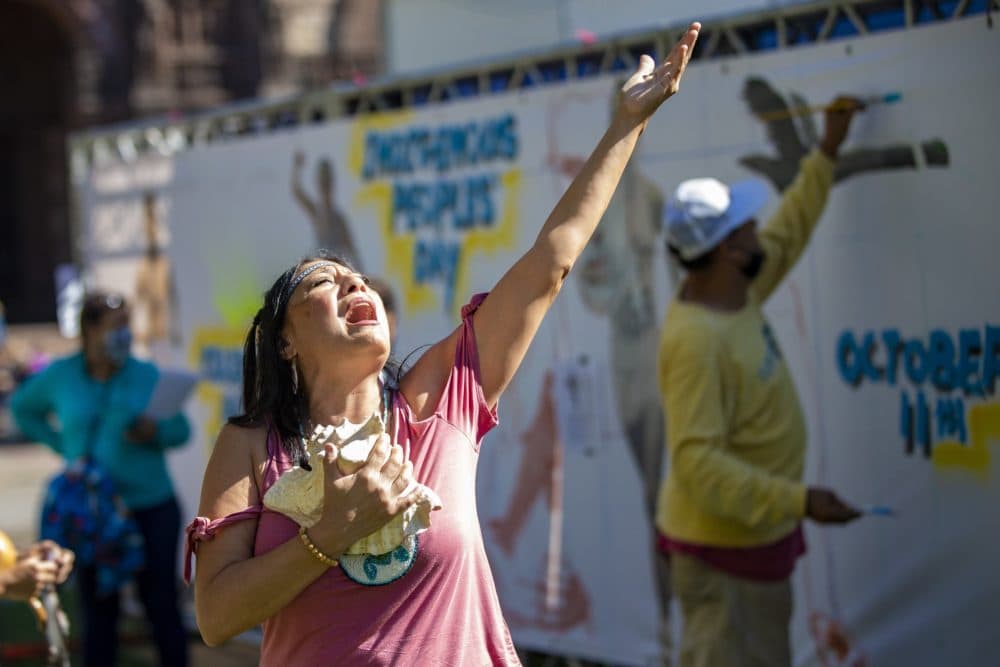 Chali’Naru Dones, co-founder of the Indigenous Peoples' Day Committee in Newton, performs a healing song as artist Robert Peters paints a mural illustrating Indigenous history in the Boston Marathon near the finish line in Copley Square. (Jesse Costa/WBUR)